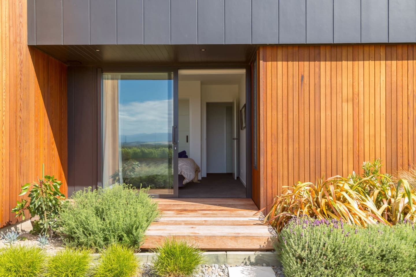 Een modern gebouw met een houten gevel, een glazen entree en een pad dat leidt naar een slaapkamer met een bed en uitzicht op de omgeving.