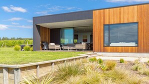 Une maison moderne dotée d’une grande façade en bois, d’un espace de détente extérieur couvert avec des chaises et une table, et offrant une vue sur un vignoble en arrière-plan.