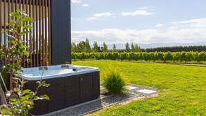 A modern building with a hot tub in a lush green field, surrounded by vineyards and mountains in the background. - 89 In The Vines, Martinborough (Martinborough)