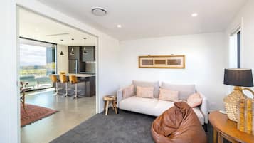 Un saló modern amb un sofà, un seient de ploma, una taula petita i una làmpada. Es veu una zona de cuina a través d'una porta oberta.