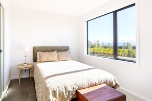 A bedroom with a large window, a bed with a headboard, a bedside table with a lamp, and a bench.