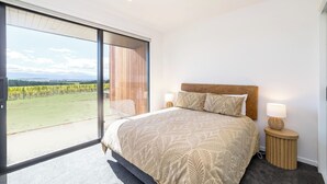 A bedroom with a large bed, two bedside lamps, and a view of a vineyard through a sliding glass door. - 89 In The Vines, Martinborough (Martinborough)