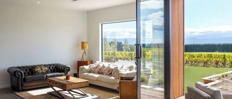 Un salon moderne avec un canapé en cuir, un canapé blanc et une table basse en bois. Une grande porte-fenêtre coulissante donne accès à un balcon offrant une vue sur un vignoble.