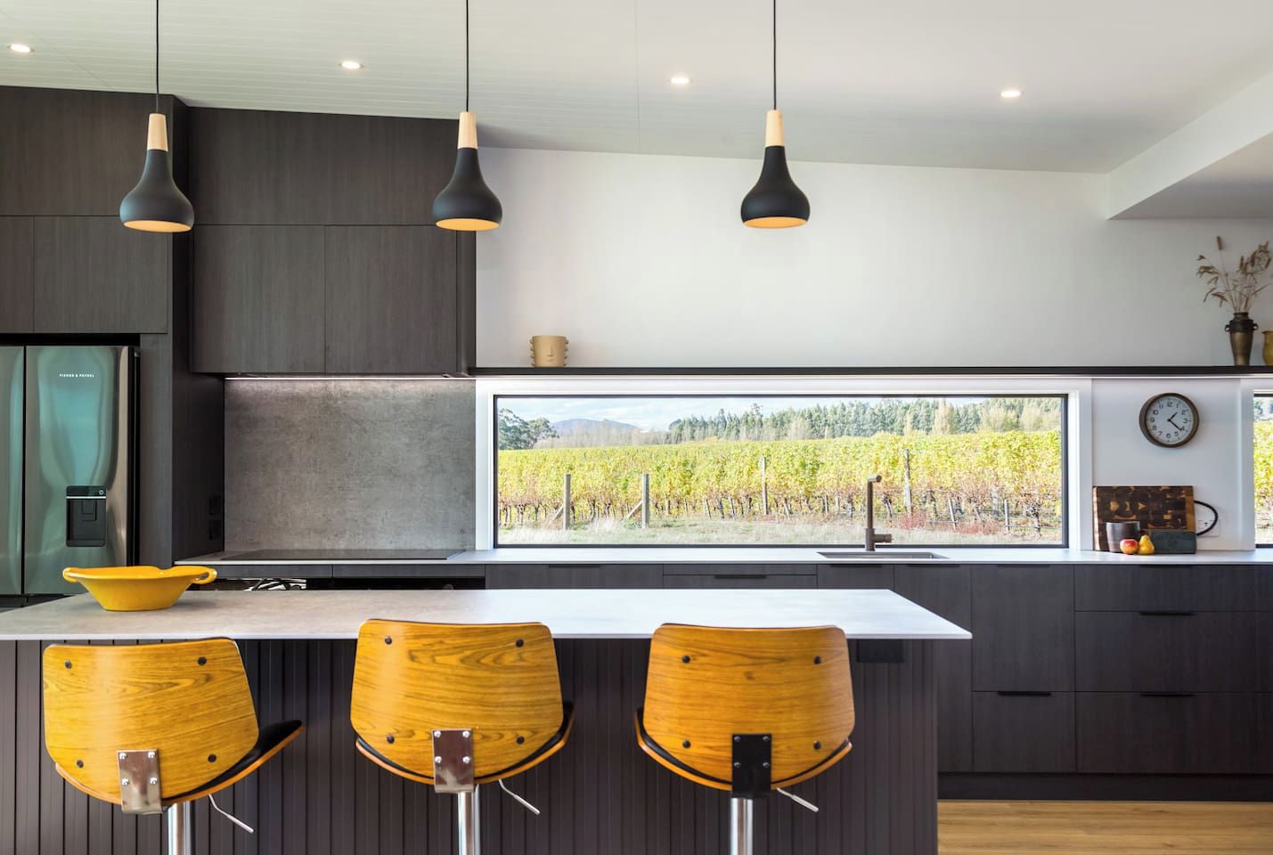 A modern kitchen with dark cabinetry, a large window with a vineyard view, and three wooden bar stools.