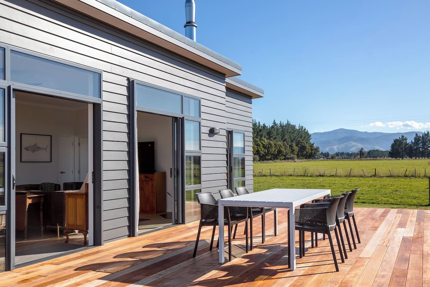 Ein modernes Haus mit einer Terrasse, einem Essbereich mit Stühlen und Blick auf ein grünes Feld und Berge.