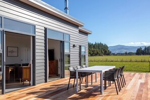 A modern house with a deck, a dining area with chairs, and a view of a green field and mountains.