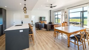 Una cocina moderna con una isla central, un comedor con mesa y sillas de madera, una sala de estar con sofá y televisor, y amplias ventanas con vistas al exterior.