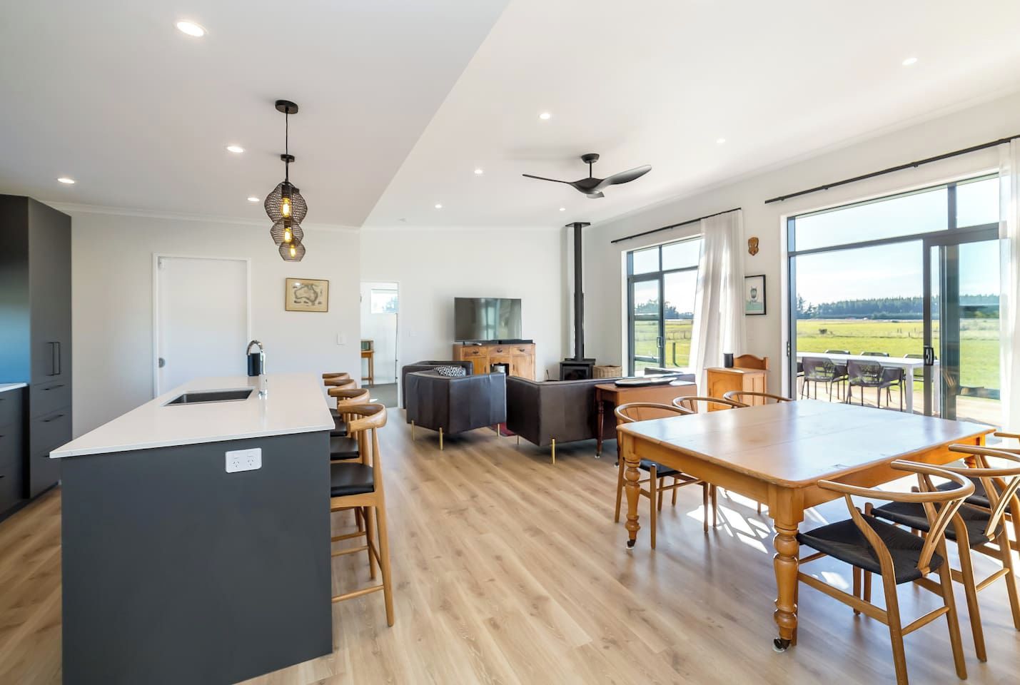 A modern kitchen with a central island, bar stools, a dining table with chairs, a fireplace, and large windows offering a view of the outdoors.