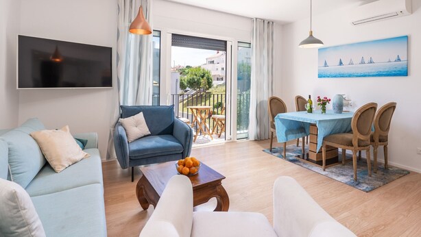 Ein modernes Wohnzimmer mit Sofa, Couchtisch mit Orangen, Essbereich mit Tisch und Stühlen, einem an der Wand montierten Fernseher und einem Balkon mit Meerblick.