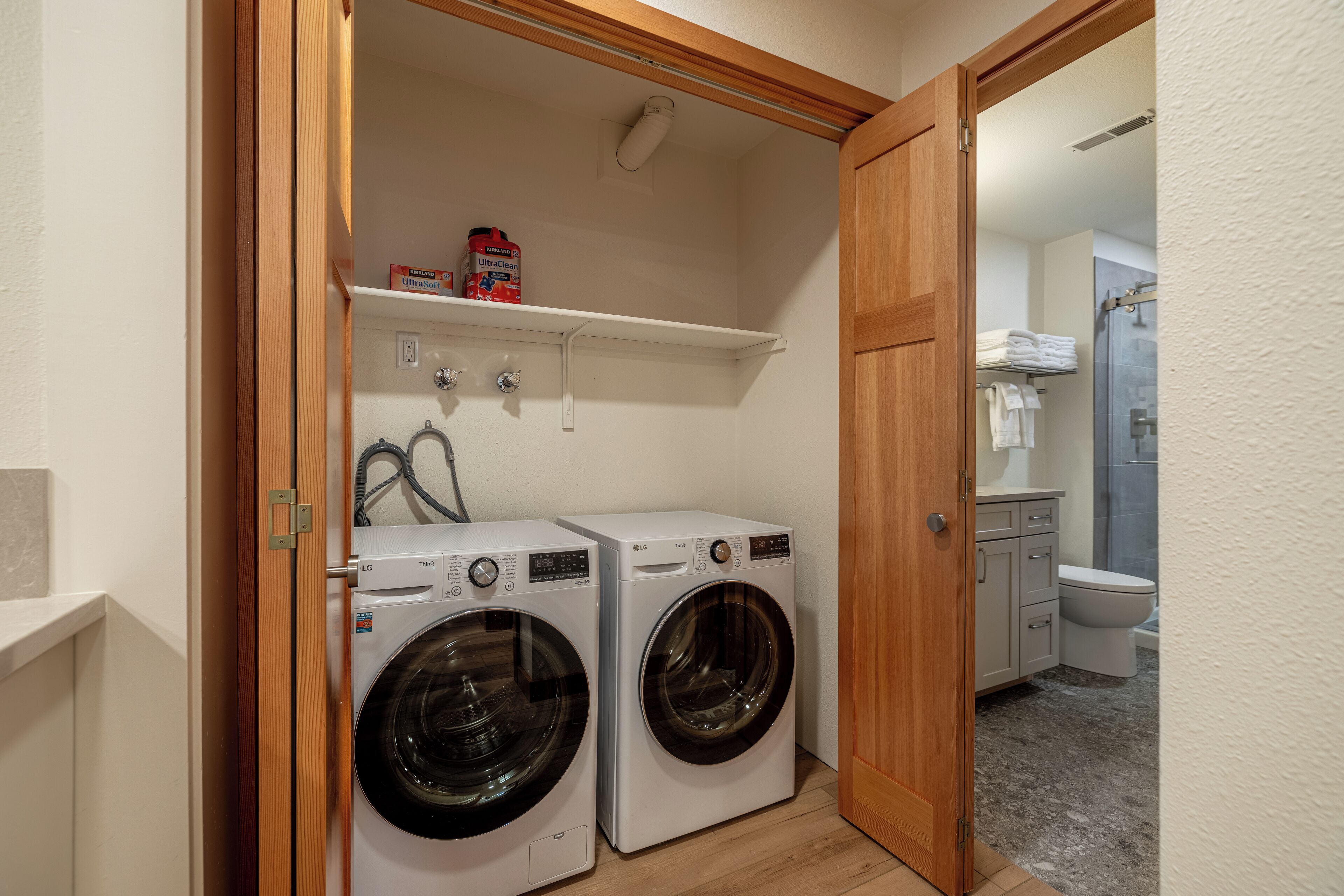 Une buanderie équipée de deux machines à laver à chargement frontal, d’une étagère contenant des produits de nettoyage et d’une porte en bois donnant accès à une salle de bain.