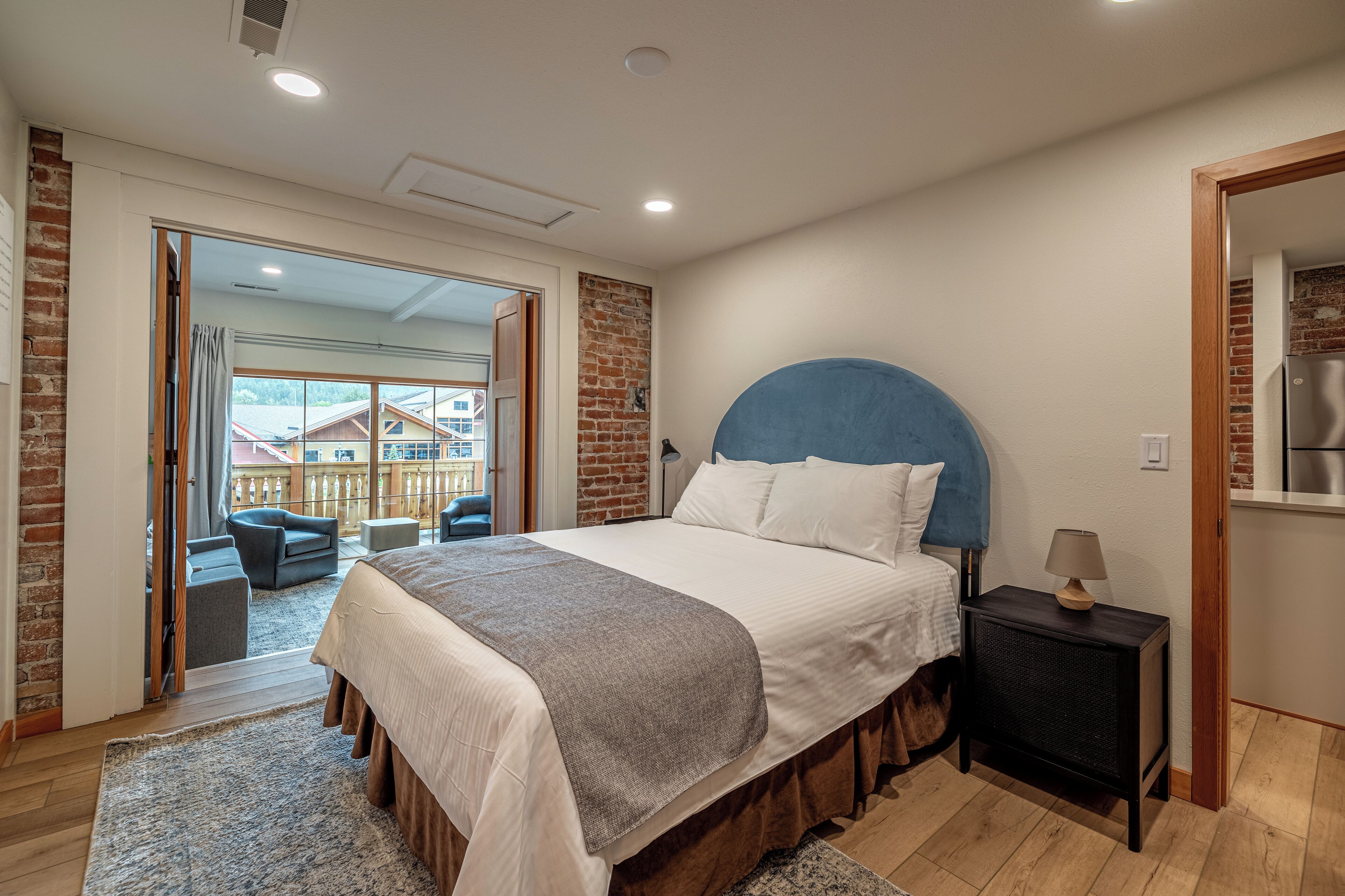 A bedroom with a large bed, a nightstand, a lamp, and a view of a balcony with outdoor furniture.