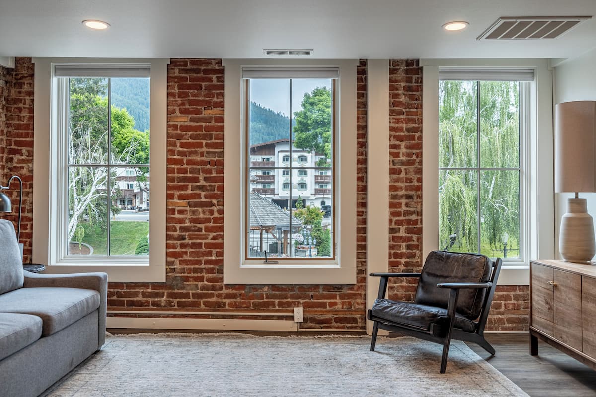 A room with a brick wall, large windows, a leather chair, and a wooden cabinet.