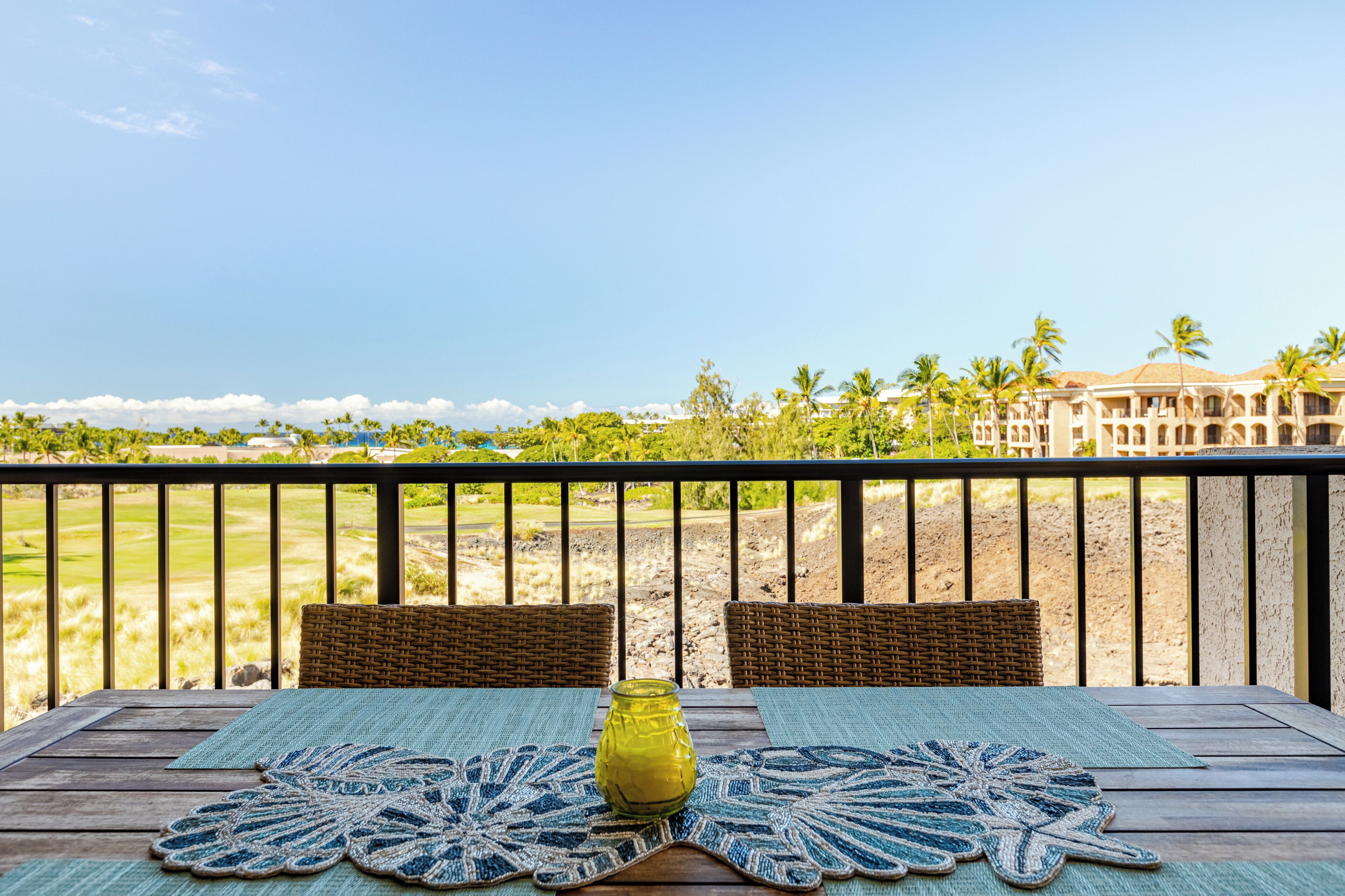 A wooden table with woven chairs and a blue and white doily, a yellow vase, and a view of a golf course and palm trees.