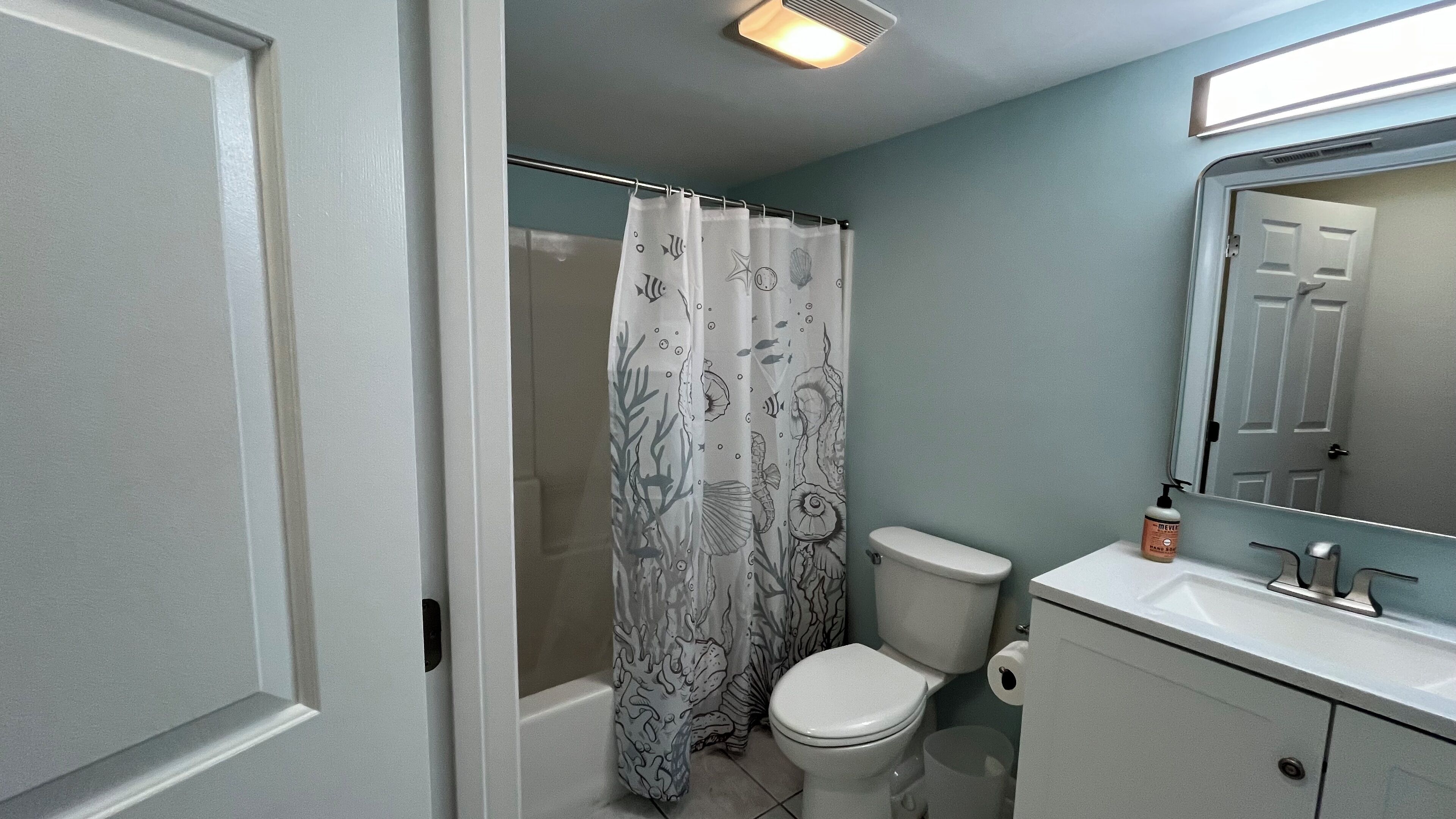 Une salle de bain avec un rideau de douche orné de motifs représentant la vie marine, des toilettes, un lavabo avec un miroir au-dessus et une porte.