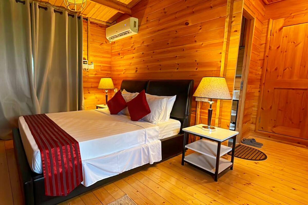 A wooden cabin room with a bed, two bedside lamps, a heater, and a window with curtains.