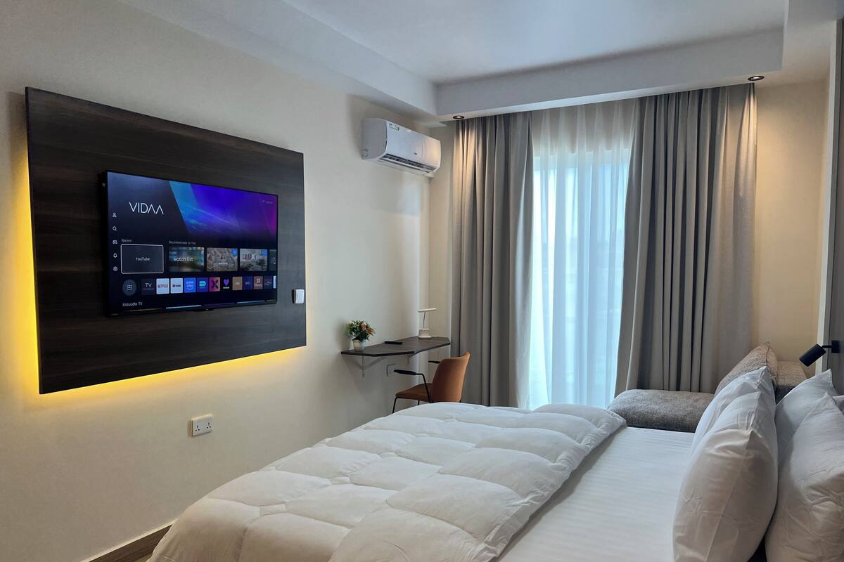 A hotel room with a bed, a TV mounted on the wall, a desk, and a window with curtains.