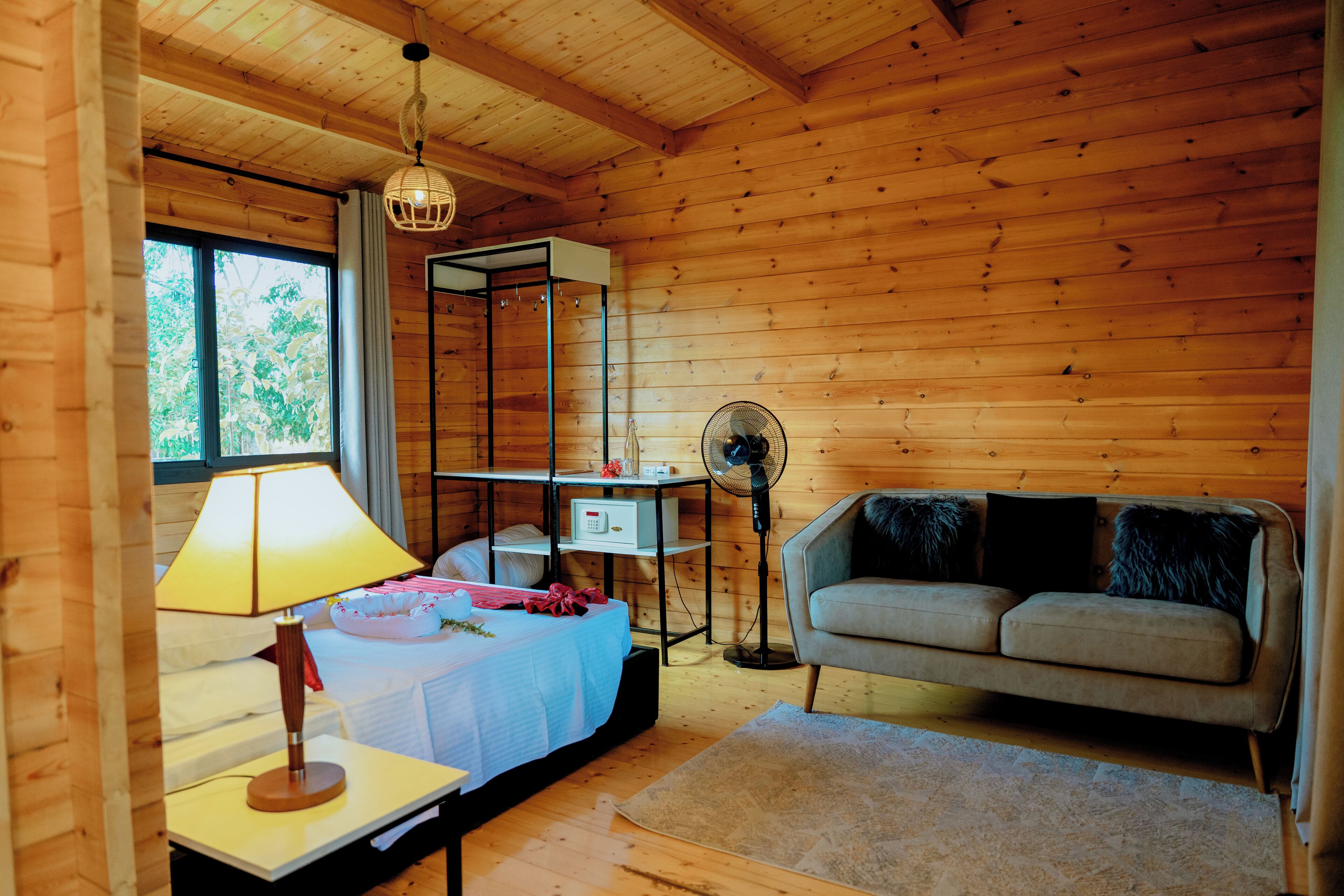 Una habitación acogedora con cama, mesita de noche con lámpara, sofá, ventilador de pie y un ventanal grande.