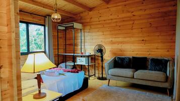 Una habitación acogedora con cama, mesita de noche con lámpara, sofá, ventilador de pie y un ventanal grande.