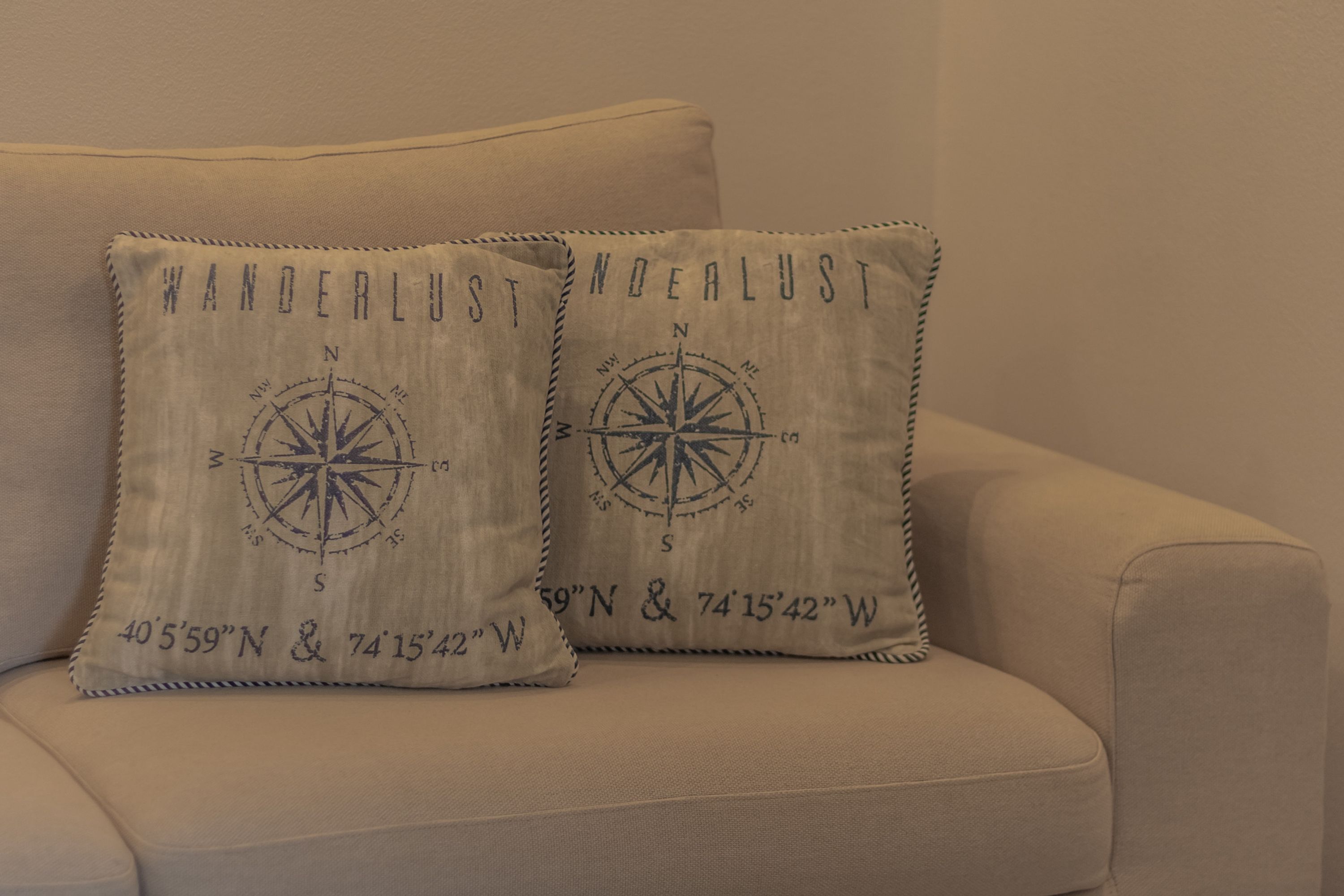 Deux coussins sur un canapé beige orné d’un motif de boussole et de coordonnées géographiques.