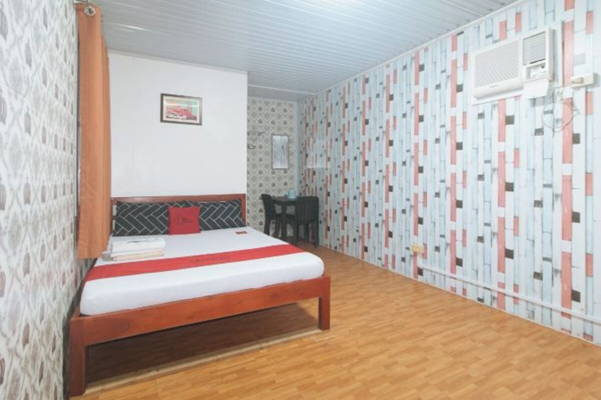 Un dormitorio con una cama de madera, una mesita pequeña y una silla. La habitación tiene papel tapiz estampado y un ventilador de techo.