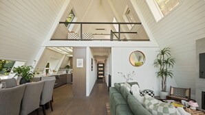 A spacious loft-style living area with a high ceiling, a dining area with chairs and a table, and a cozy seating area with a sofa and cushions. - The Gnome Gully - A-Frame w/ Sound Views (Olympia)