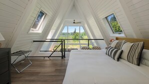A bedroom with a sloped ceiling, a bed with striped pillows, a ceiling fan, and a view of greenery through large windows. - The Gnome Gully - A-Frame w/ Sound Views (Olympia)