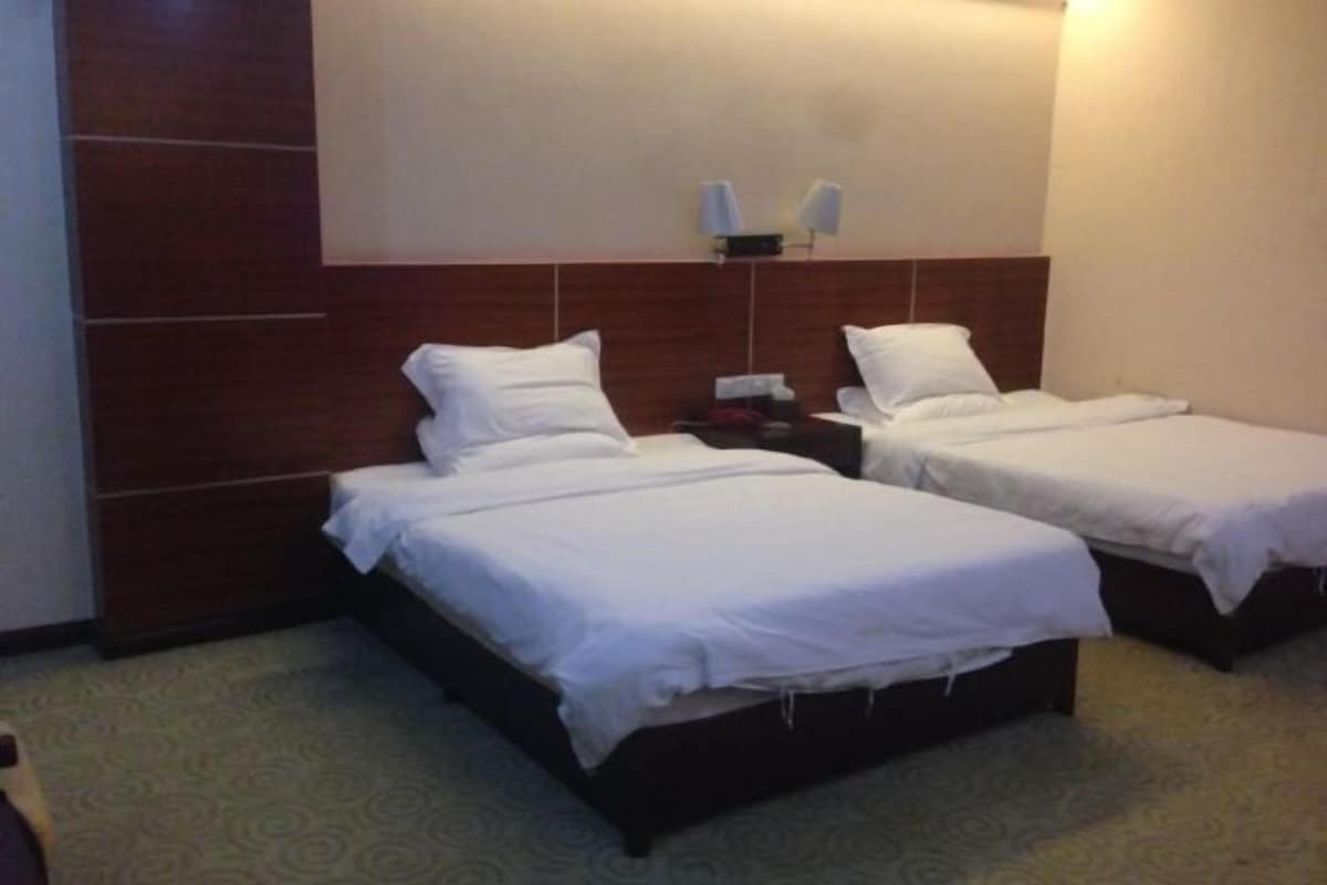A hotel room with two single beds, a wooden headboard, a small bedside table with a lamp, and a wall-mounted light fixture.