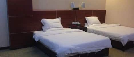 A hotel room with two single beds, a wooden headboard, a small bedside table with a lamp, and a wall-mounted light fixture.