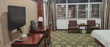 A hotel room with a bed, a desk with a TV, two chairs, a window with curtains, and a patterned carpet.