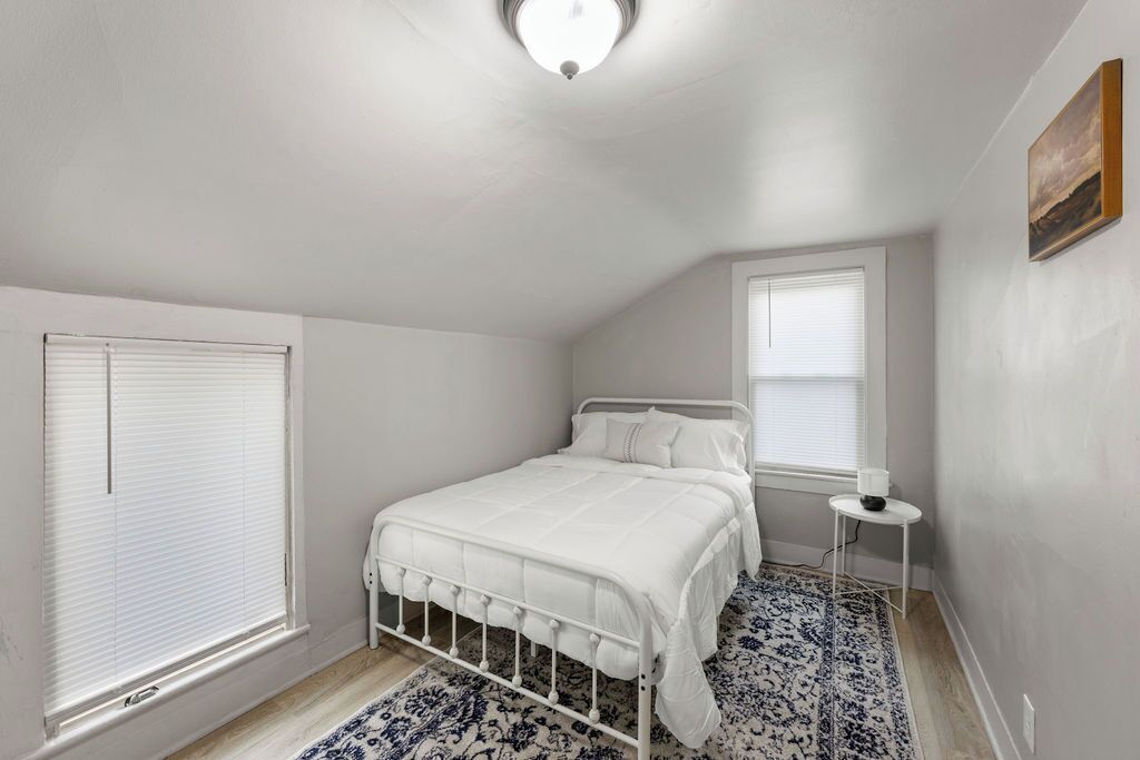 Un dormitorio con una cama blanca, una mesita de noche pequeña, una lámpara de pie y una alfombra estampada.