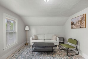 Habitació petita i ben il·luminada amb un sofà blanc, una cadira verda, una taula de cafè negra i una làmpada de peu. Hi ha un quadre a la paret i una finestra amb persianes a l'esquerra.