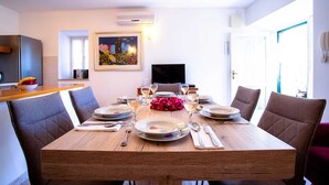 A dining area with a wooden table set for four, surrounded by chairs. There is a painting on the wall, a television, and a kitchen area visible in the background. - Charming 3-BR Villa for 6 with private pool, close to the sea (Sumartin)
