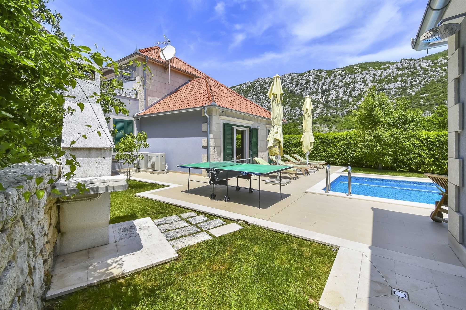 A pool area with a ping pong table, lounge chairs, and a small building with a red-tiled roof.