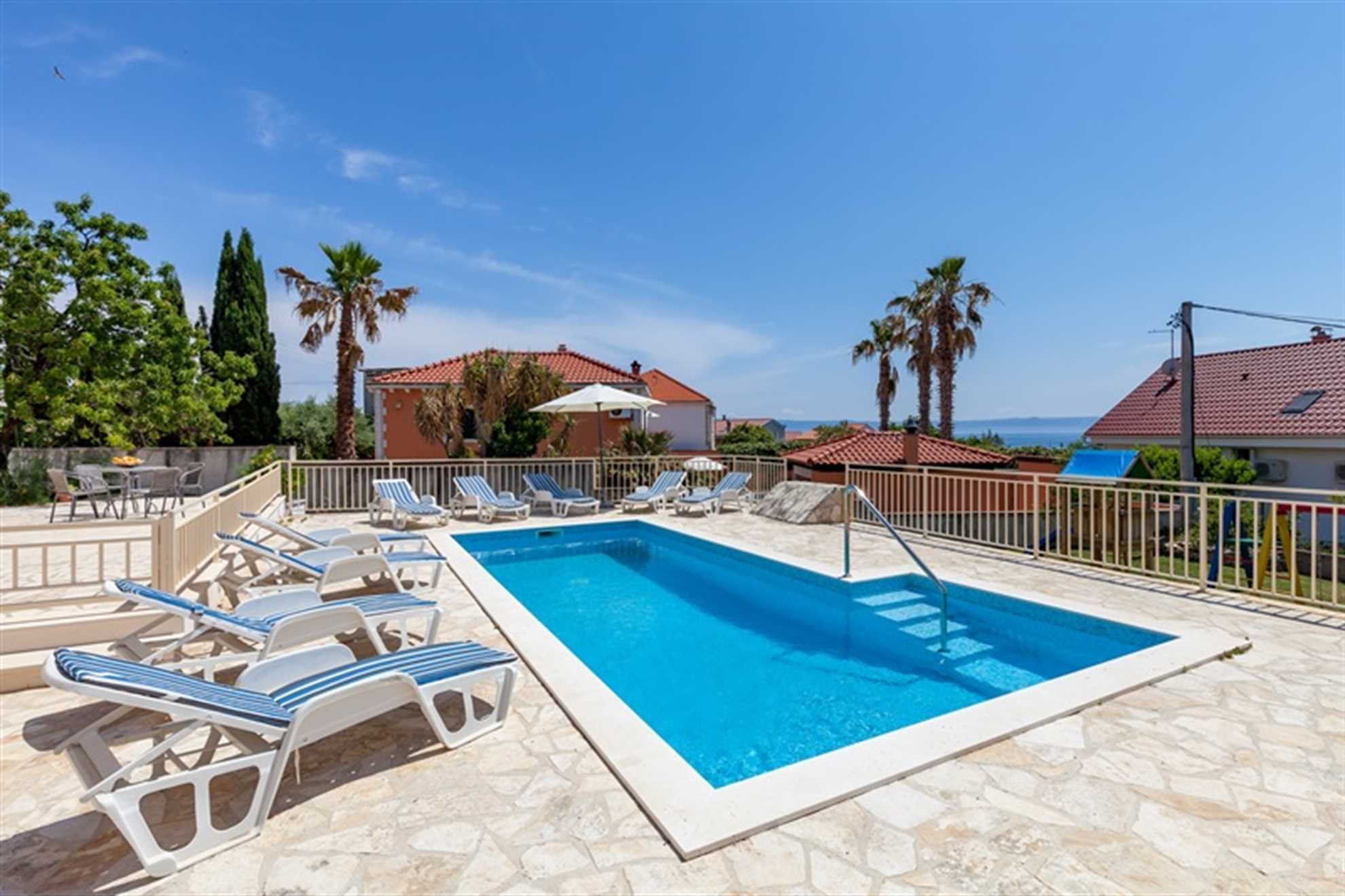 Une piscine avec des chaises longues, une terrasse en pierre et une vue sur la mer et les bâtiments.