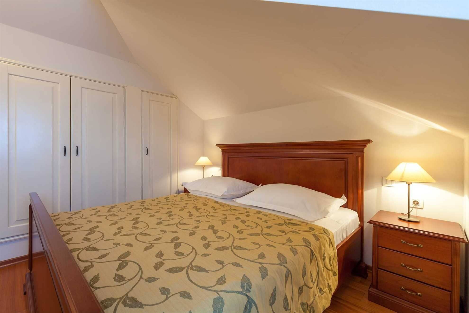 A bedroom with a bed, two bedside tables with lamps, a wardrobe, and a wooden dresser.