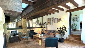 Ein gemütliches Wohnzimmer mit Steinwand, Holzbalken, einer Couch, Sesseln, einem Couchtisch und einer Treppe, die zu einer weiteren Ebene führt.