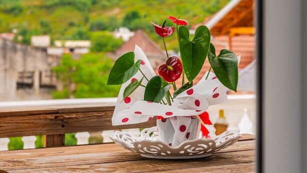 Una mesa de madera con un jarrón blanco que contiene flores rojas y hojas verdes, frente a un paisaje verde y un cielo nublado.