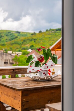 Une table en bois sur laquelle est posé un vase blanc contenant des fleurs rouges et des feuilles vertes, le tout se détachant sur un paysage verdoyant et un ciel nuageux.