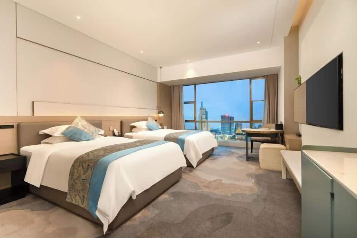 A hotel room with two beds, a desk, a chair, a TV, and a large window with a city view.
