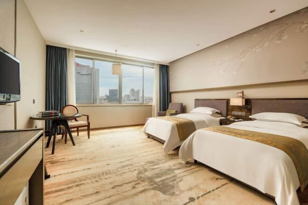 A hotel room with two beds, a desk, a chair, a TV, and a large window with a city view.