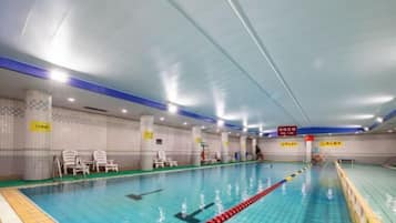 An indoor swimming pool with clear lane dividers, white tiled walls, and several lounge chairs along the sides.