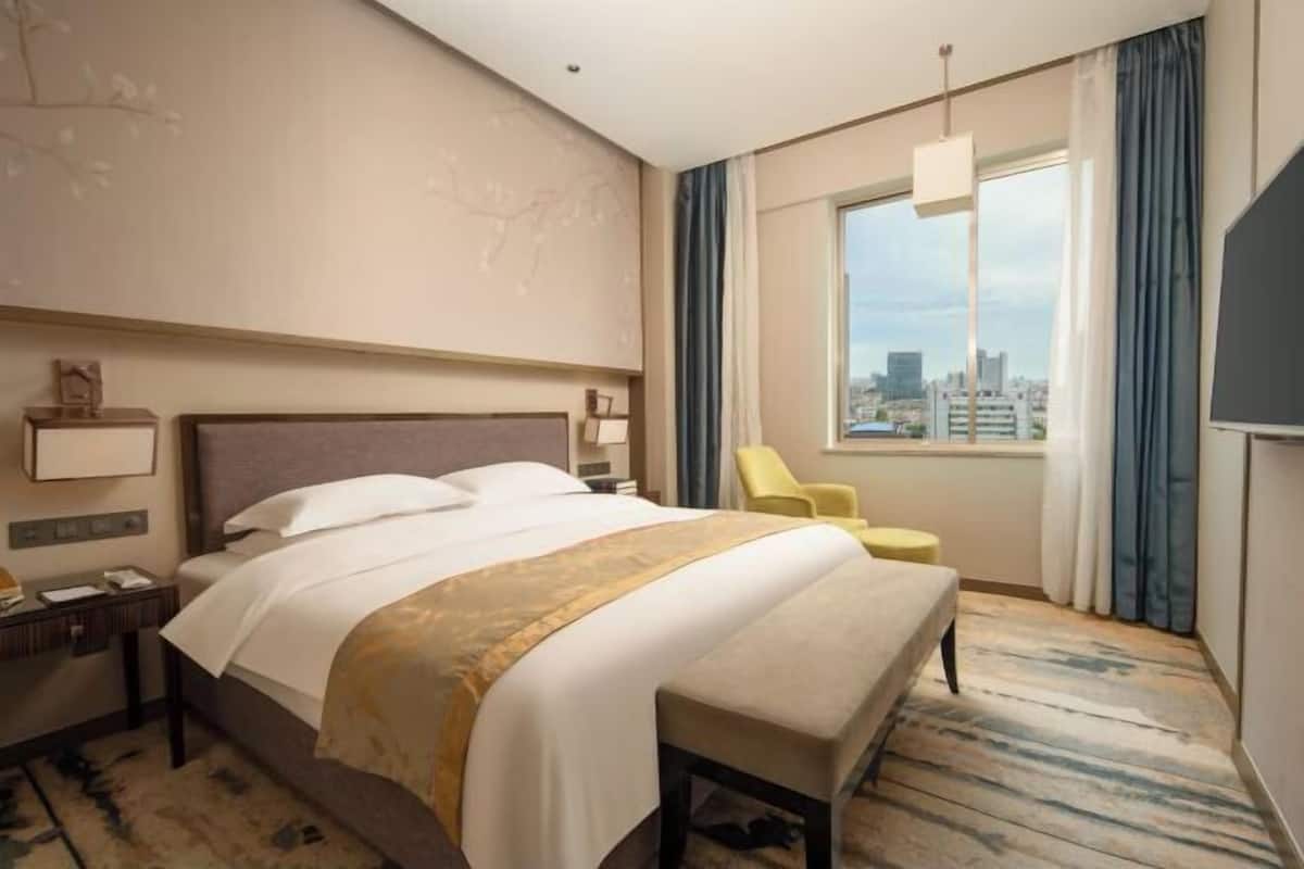 A hotel room with a large bed, a bench, a chair, a desk, and a window with a city view.