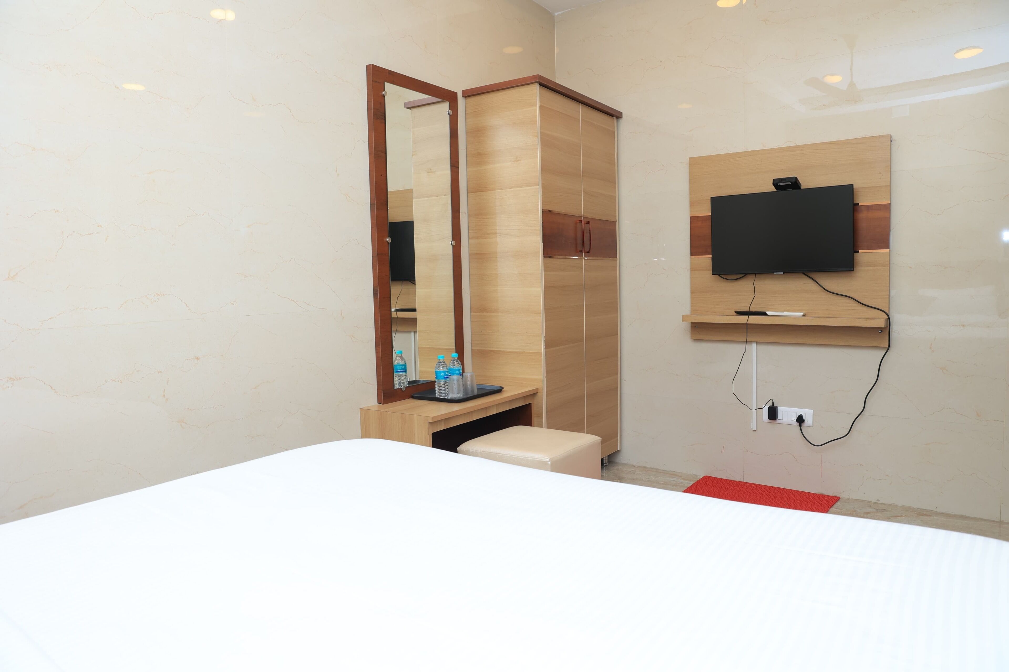 Una habitación de hotel moderna con una cama, un armario de madera, un espejo, un televisor montado en la pared y una pequeña mesa con una botella de agua.
