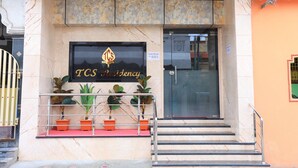 A TCS Residency building entrance with a marble facade, potted plants, and a contact sign. - TCS Residency (Kanchipuram)