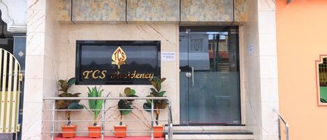 A TCS Residency building entrance with a marble facade, potted plants, and a contact sign.