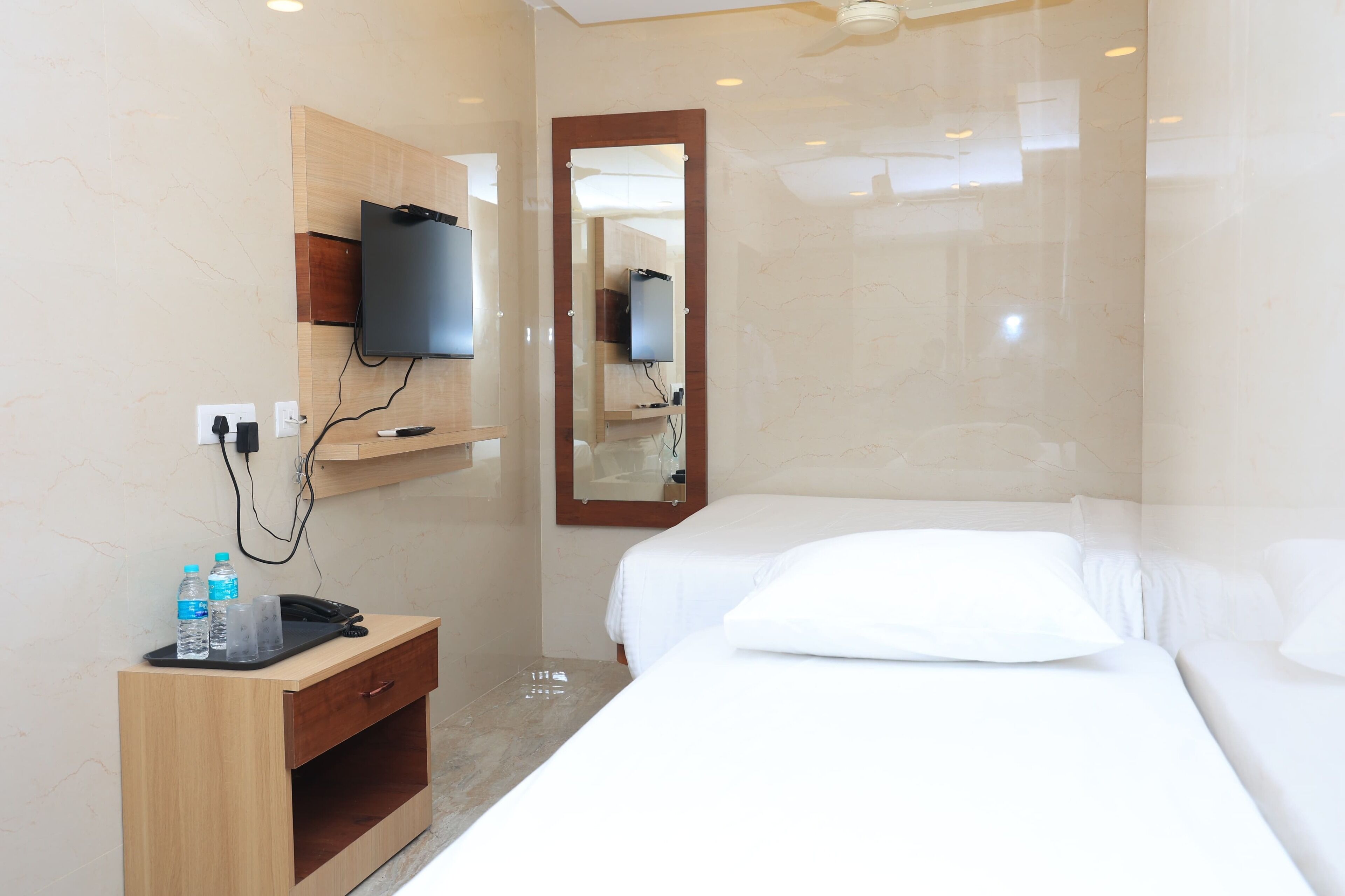 Habitación de hotel con cama, mesita de noche con teléfono y botellas de agua, televisor montado en la pared y espejo.