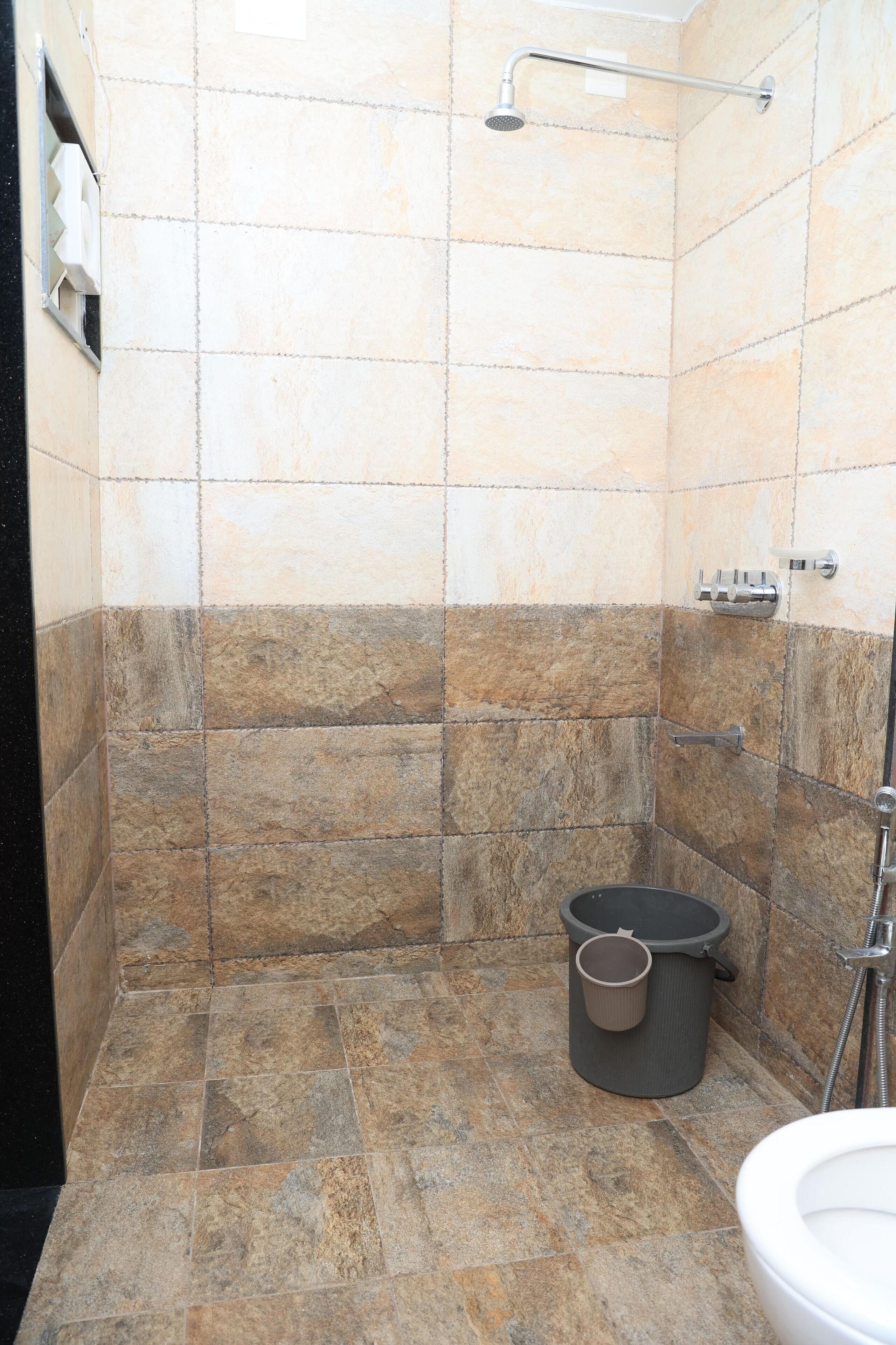 A bathroom with beige and brown tiles, a shower, a toilet, and a trash bin.