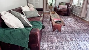 Amplia sala de estar con un sofá de cuero marrón grande, un sill ón a juego y una alfombra estampada. Hay una mesita de centro, una lámpara de pie y una planta en maceta.
