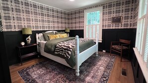 Un dormitorio con cama, mesitas de noche, una silla, una ventana y papel tapiz estampado.