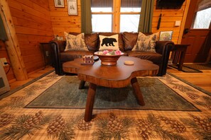 A cozy cabin interior with a wooden floor, a brown leather sofa with a bear pillow, a wooden coffee table, and a pine-patterned rug. - Cozy Log Cabin, Sleeps 6, Pet Friendly! (Norway)